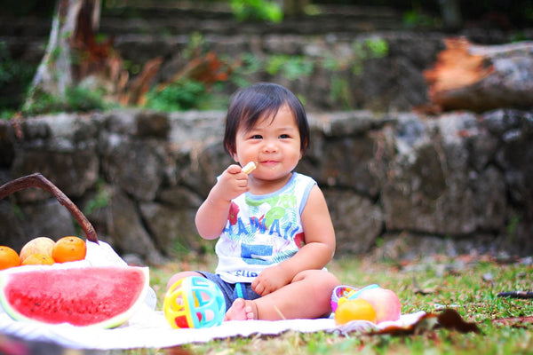 Baby mit Spielzeug auf Picknickdecke und isst Snacks. Neben dem Baby ist ein Obstkorb und angeschnittene Melone
